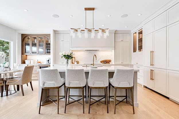 Modern kitchen with white cabinets, a large island with five cushioned stools, gold accents, pendant lights, and a breakfast nook by the window. Marble backsplash and wood floors add elegance.