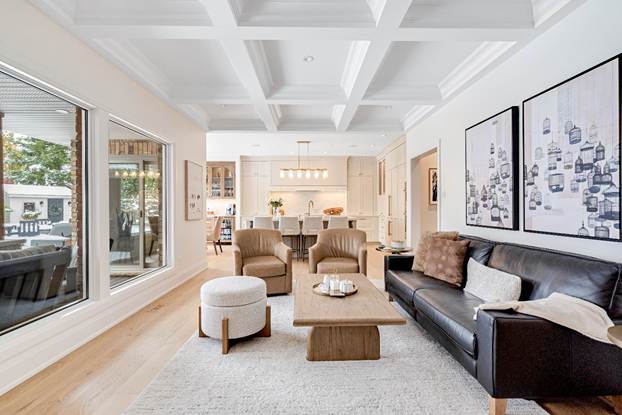 Modern living room with large windows, a black leather sofa, two beige chairs, a wooden coffee table, and wall art. The space opens to a bright kitchen with white cabinetry and barstools at the island.