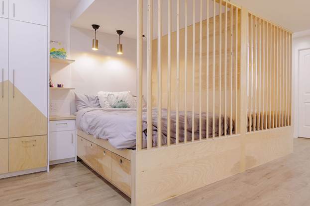 Modern bedroom with a wooden slatted partition, built-in storage drawers under the bed, white and light wood cabinetry, and soft lighting from two pendant lamps.