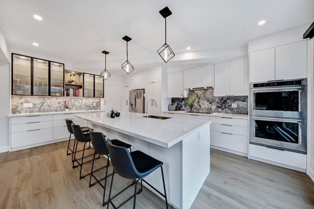 Modern kitchen with white cabinets, marble countertops, three black barstools, stainless steel appliances, pendant lights, and wood flooring. Black-framed glass cabinets and a large island complete the design.