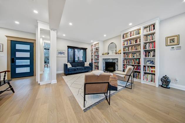 Modern living room with wooden floors, built-in bookshelves, a dark blue sofa, two beige chairs, a fireplace, and a geometric rug. A blue front door and large window are visible in the bright, open space.