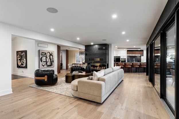 Modern open-concept living room with light wood flooring, beige sofa, black accent chairs, abstract wall art, a central fireplace, and a kitchen with bar seating in the background. Floor-to-ceiling windows on the right.