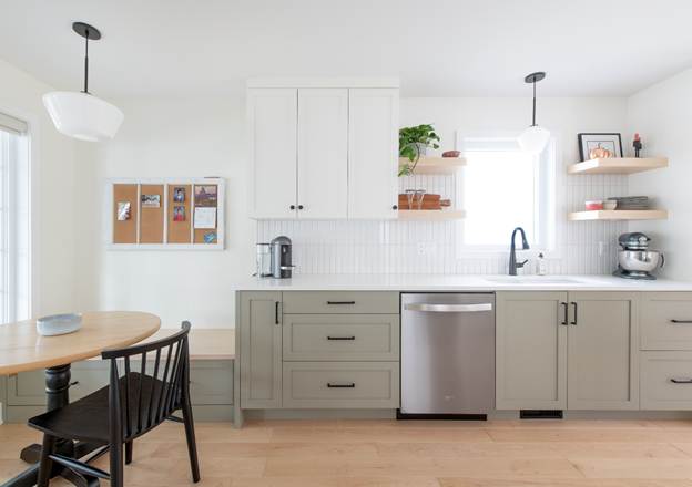 Modern kitchen with light green cabinets, white backsplash, stainless steel dishwasher, sink, open shelves with plants and dishes, a coffee maker, and a small dining table with black chairs.
