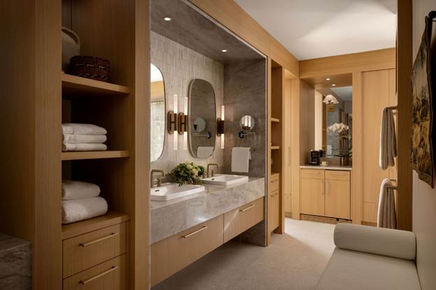 Modern bathroom with double sinks, large mirrors, built-in shelves with towels, wooden cabinets, and soft lighting. A cushioned bench sits near the vanity, and a coffee station is visible in the background.