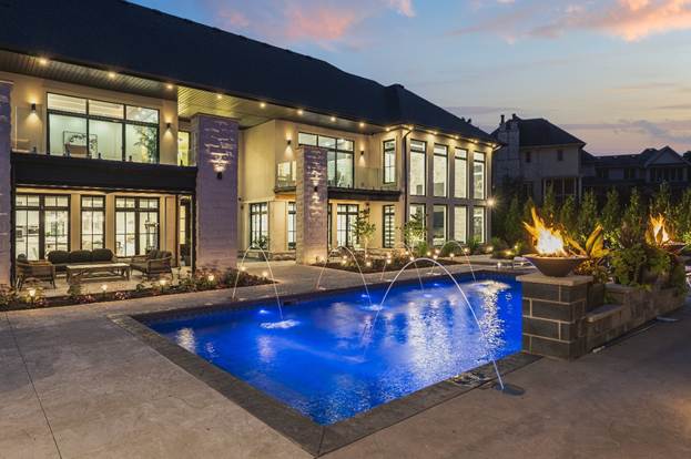 A luxurious modern house at dusk features large windows, multiple patios with seating, a lit swimming pool with water fountains, and decorative fire pits around the poolside.