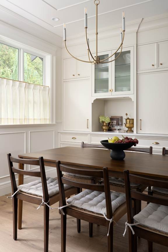 A dining room with a wooden table, six cushioned chairs, a fruit bowl centerpiece, white cabinetry, brass hardware, a window with a sheer curtain, and a modern brass chandelier above.