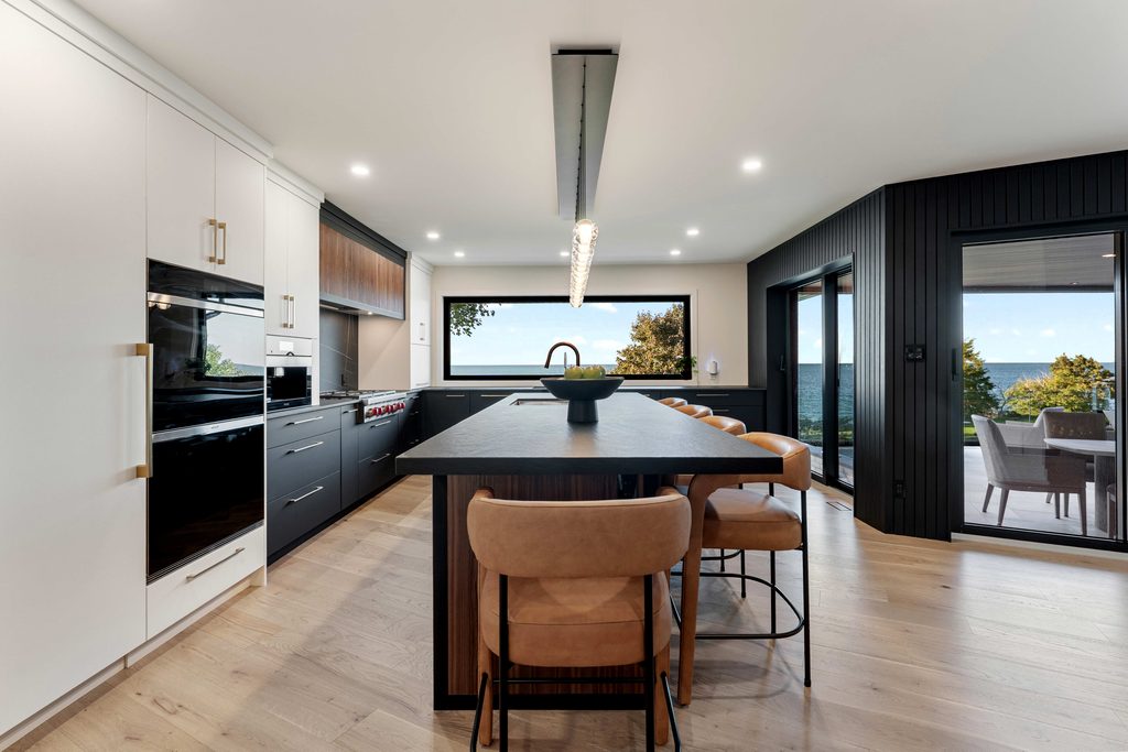 Modern kitchen with black and white cabinetry, a large island with tan chairs, light wood floors, and large windows showing trees and a view of water outside. A dining table is visible through glass doors.