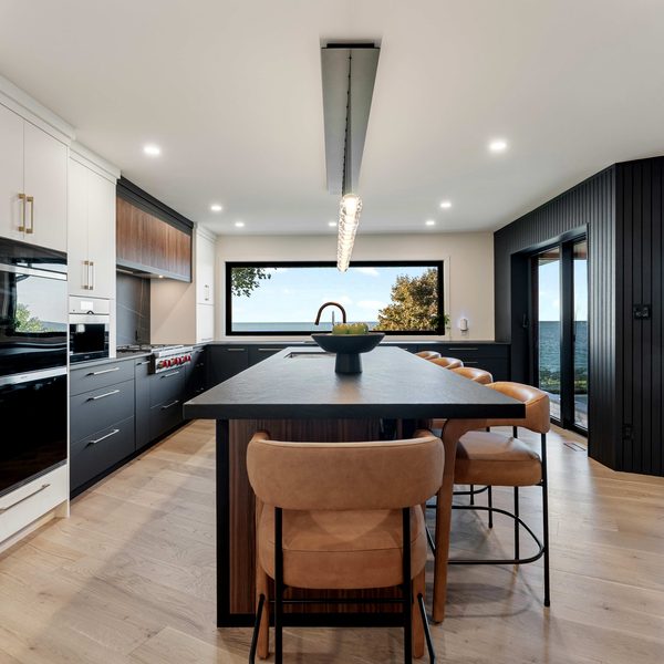 Modern kitchen with black and white cabinetry, a large island with tan chairs, light wood floors, and large windows showing trees and a view of water outside. A dining table is visible through glass doors.