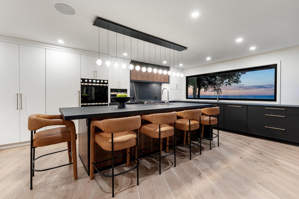 Modern kitchen with a large island, five tan barstools, pendant lighting, white cabinets, black countertops, wood accents, and a large window showing a sunset view over water.