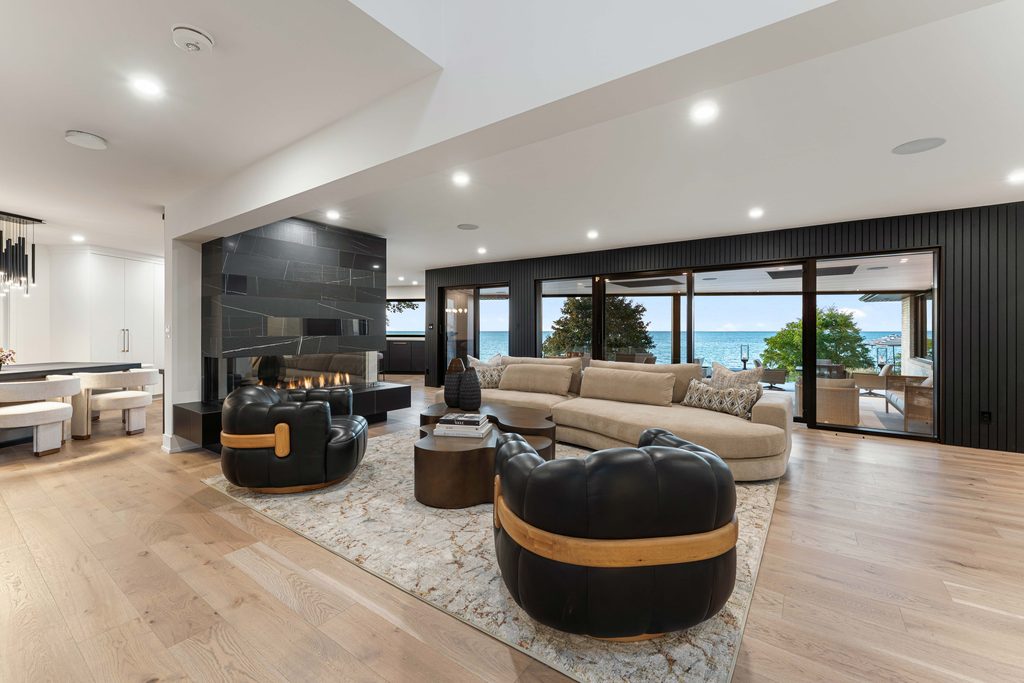 Modern living room with black sofas, a beige sectional, a fireplace, large windows, and a view of a patio and water outside. Light wood floors and recessed lighting create an open, airy atmosphere.
