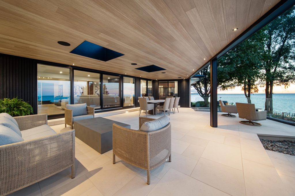 Modern outdoor patio with cushioned chairs, a dining area, and large glass doors overlooking a serene lake at sunset, surrounded by trees and minimalistic decor.