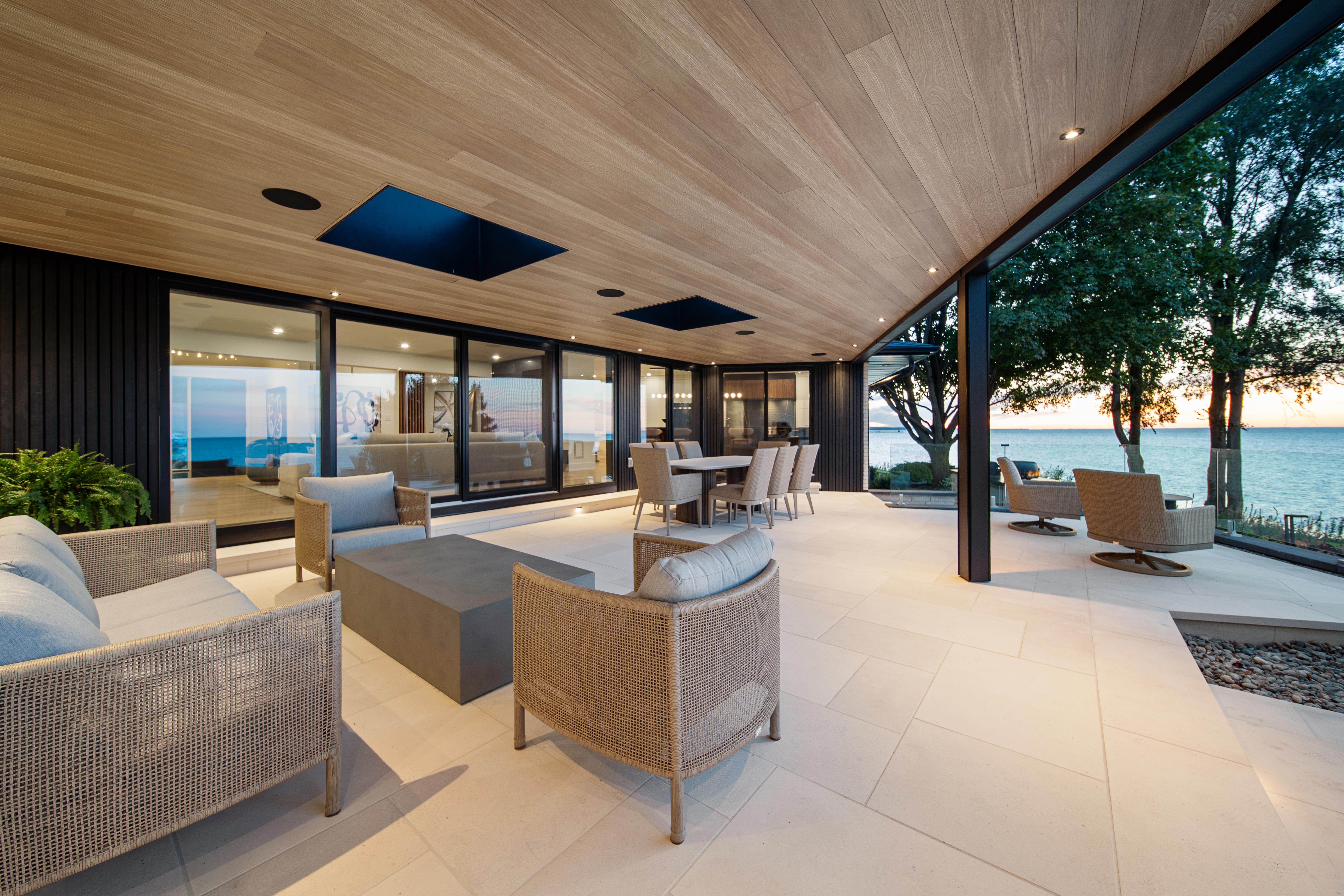 Modern outdoor patio with cushioned chairs, a dining area, and large glass doors overlooking a serene lake at sunset, surrounded by trees and minimalistic decor.