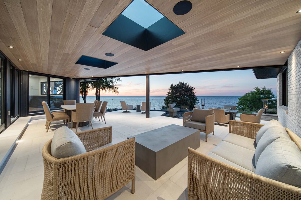 Modern covered patio with wicker furniture, a large coffee table, dining area, and open view of the ocean at sunset. The ceiling has skylights, and the space overlooks a scenic, tranquil waterfront landscape.