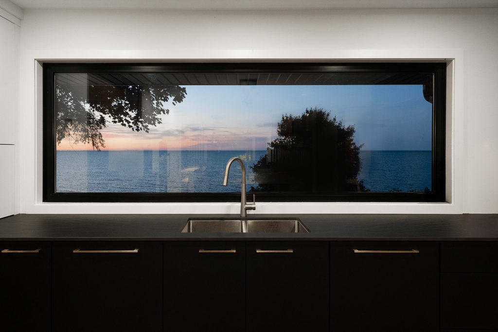 A large kitchen window frames a sunset view over a calm lake, with silhouettes of trees and a modern sink in the foreground.